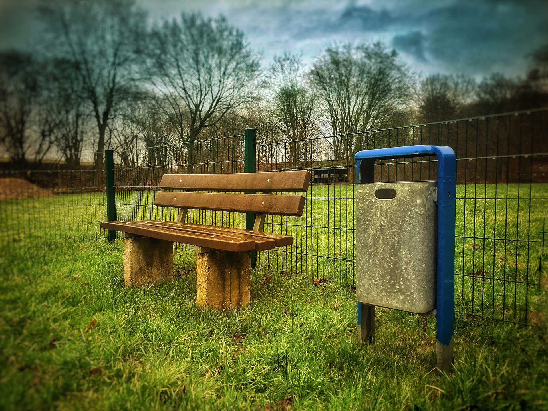 Ein blauer Mülleimer steht neben einer Sitzbank auf einer grünen Wiese. Dahinter ist ein Zaun zu sehen.