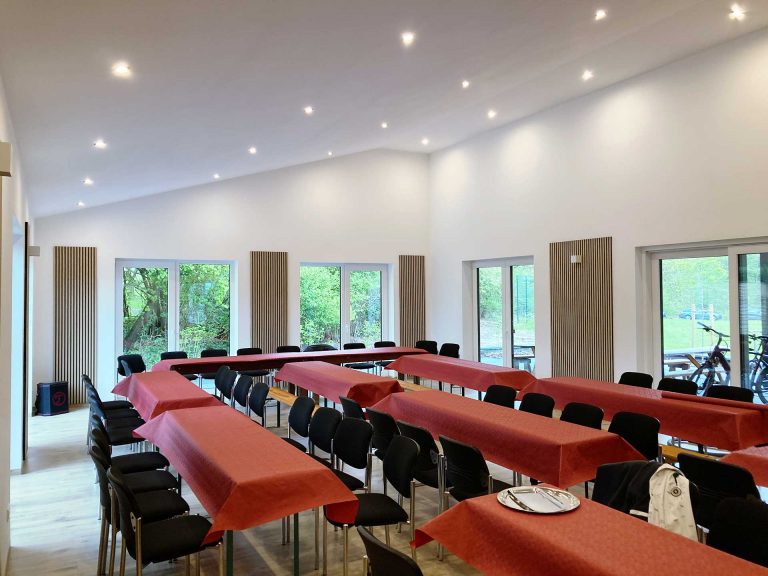 Der Saal des Selbecker Dorfgemeinschaftshauses inkl. Tischen und Bestuhlung. Auf den Tischen liegen rote Tischdecken.